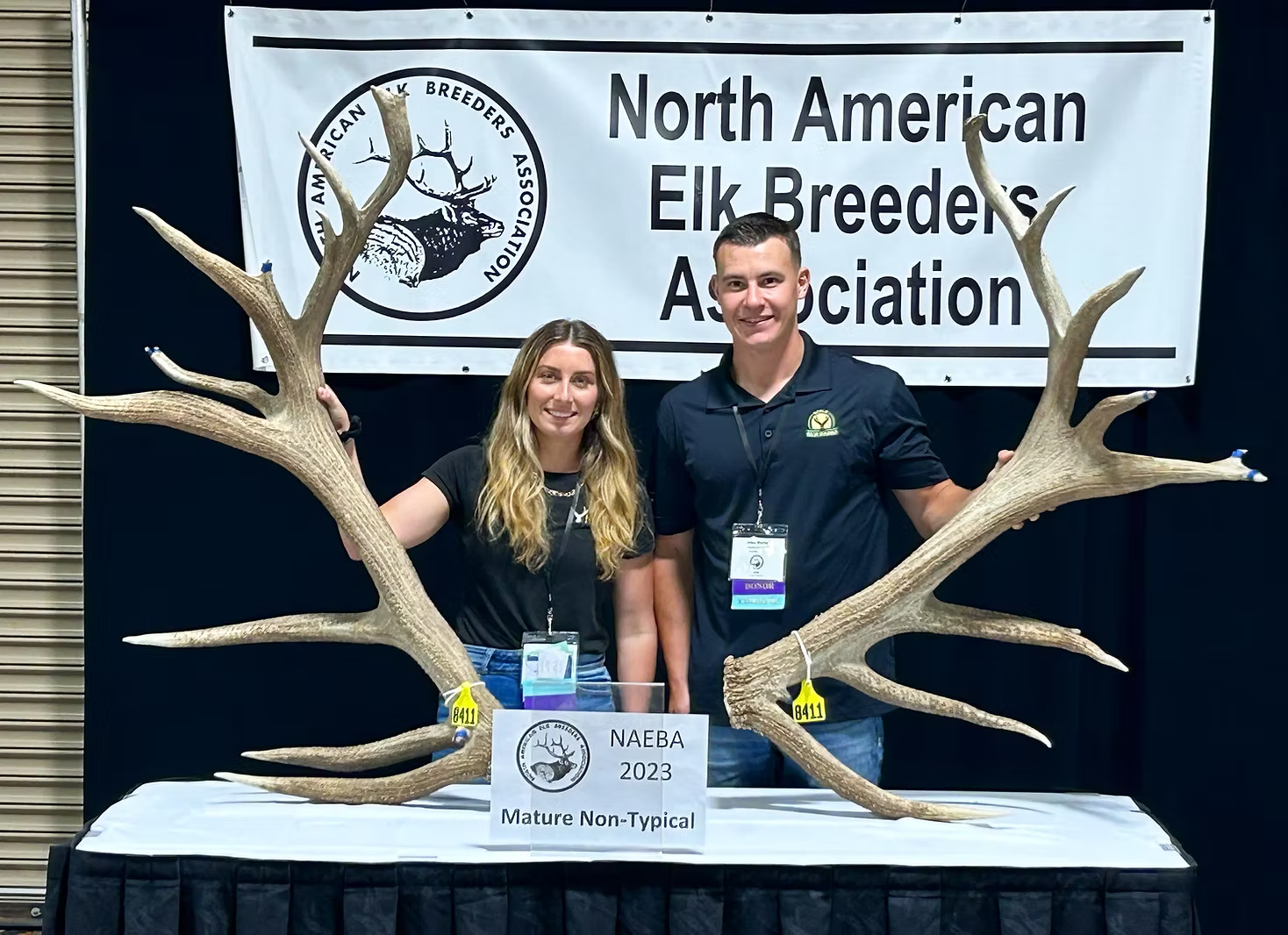 Two people standing behind a table with large elk antlers, NAEBA banner in the background