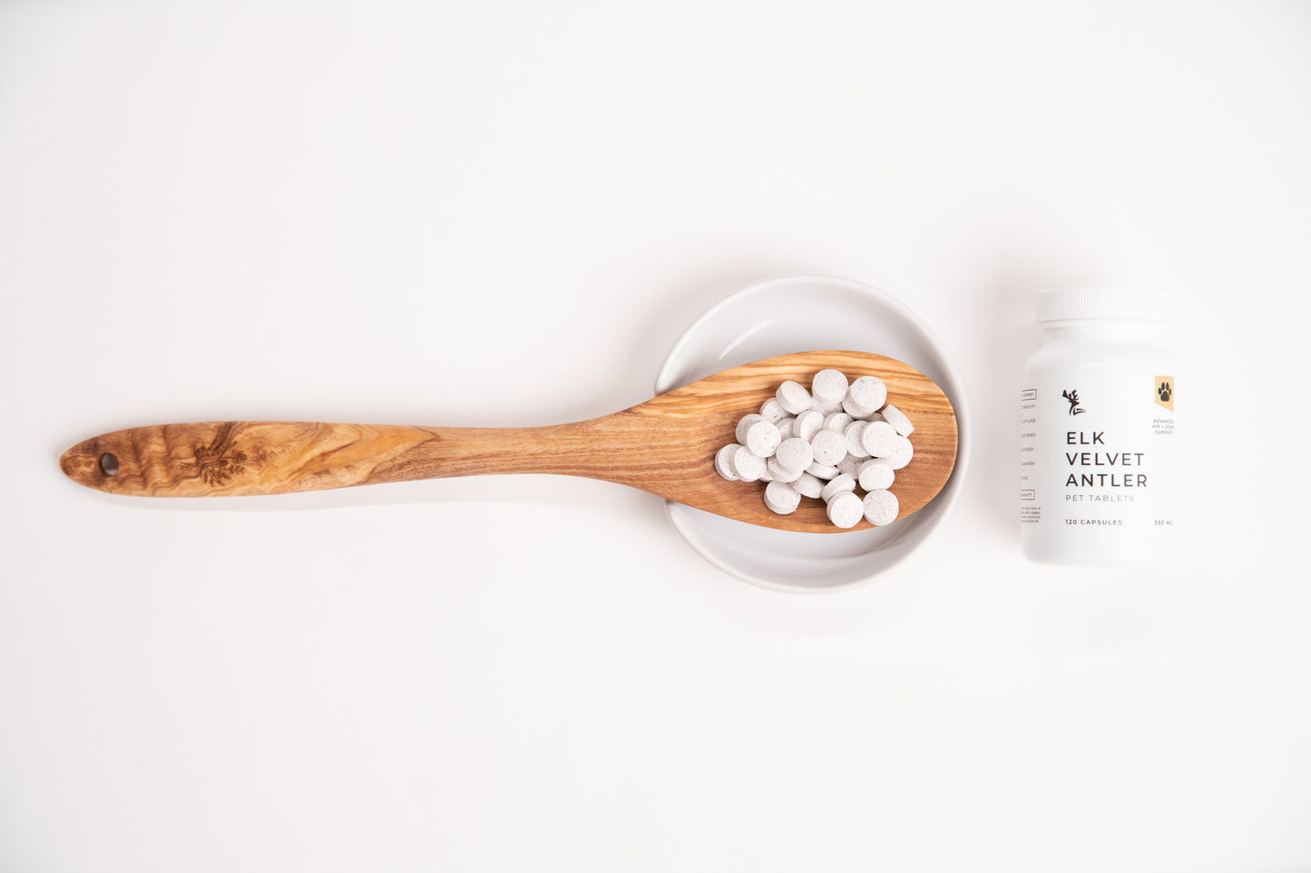 Wooden spoon with white capsules on a white background, next to a container labeled 'Elk Velvet Antler Pet Tablets'.
