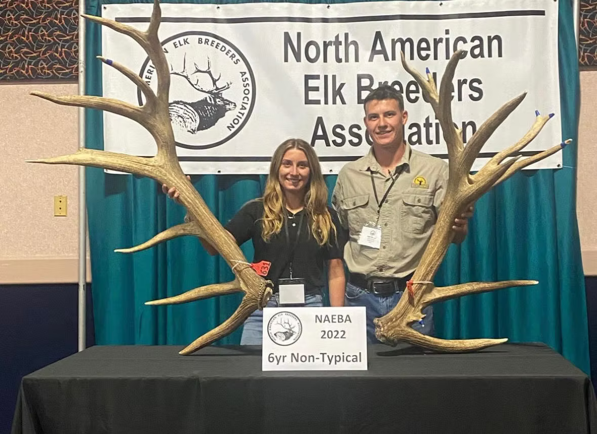 Two people holding elk antlers with a North American Elk Breeders Association banner in the background.